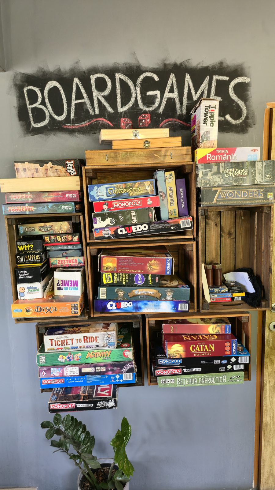 Peretele MANCAVE cu lăzi de lemn pline de boardgames, sub inscripția scrisă cu cretă
